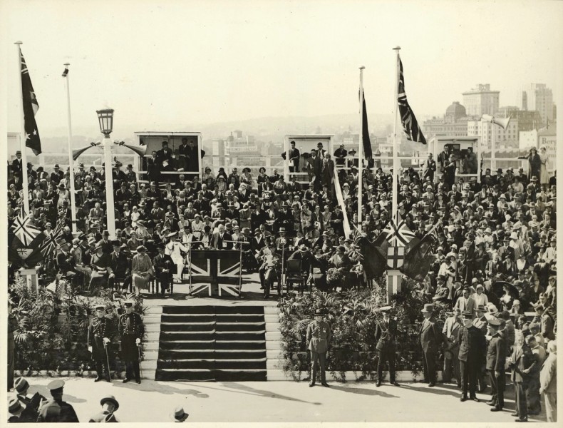 A crowd watches a man speak from a podium that is decorated with the union jack to