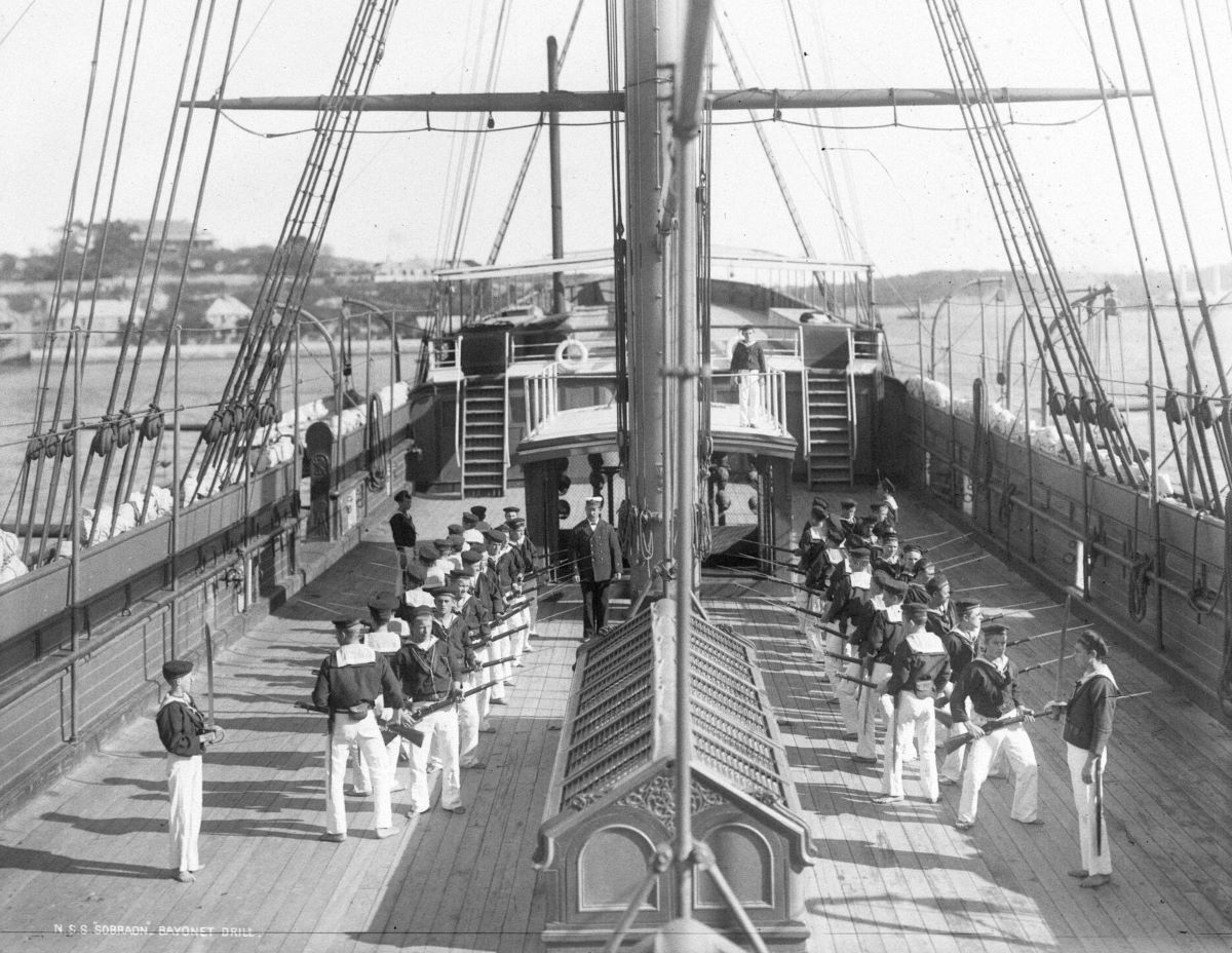 Boys in nautical school uniforms line up on the deck of a ship holding bayonets