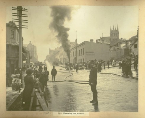 Onlookers stand behind barriers watching the streets being cleaned with fire hoses. Policemen stand on the road in front of the crowds