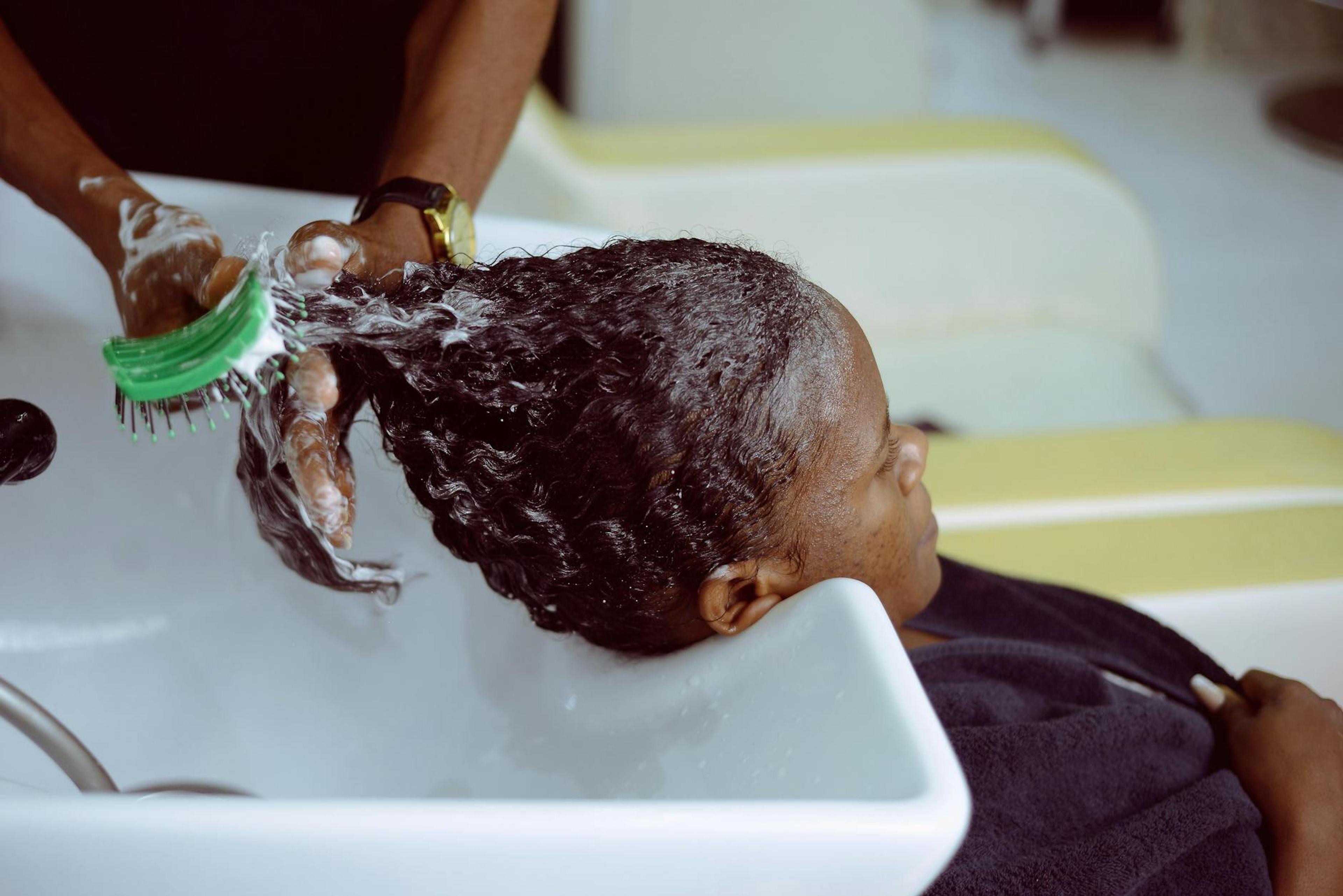 A Black woman with beautiful natural hair, smiling while applying a gentle cleanser to her scalp, showcasing a healthy hair care routine in a well-lit bathroom setting.