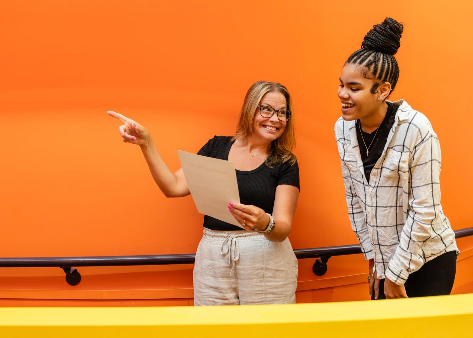 Two women stand in front of an orange wall. They book look at a piece of paper one woman is holding. She points in one direction.