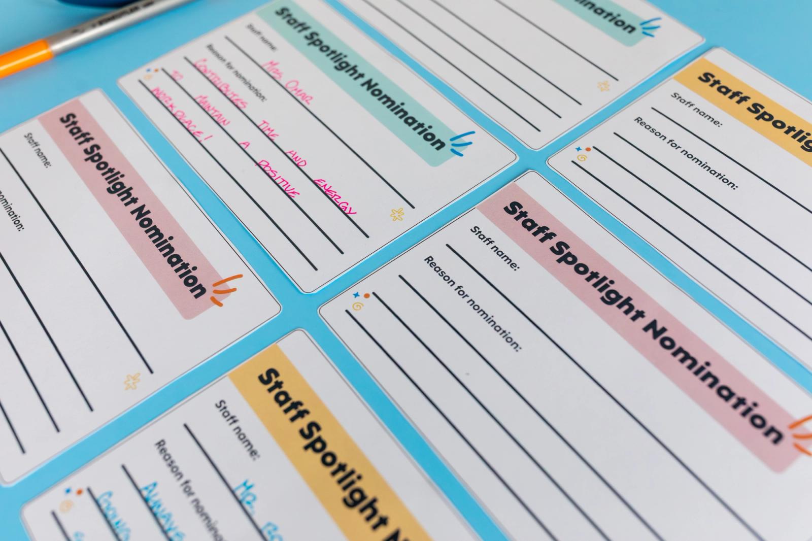 Sheets of paper lay on a light blue background. A bin with notecards and a pen in it lays on top of the paper. A pair of scissors is next to the bin.
