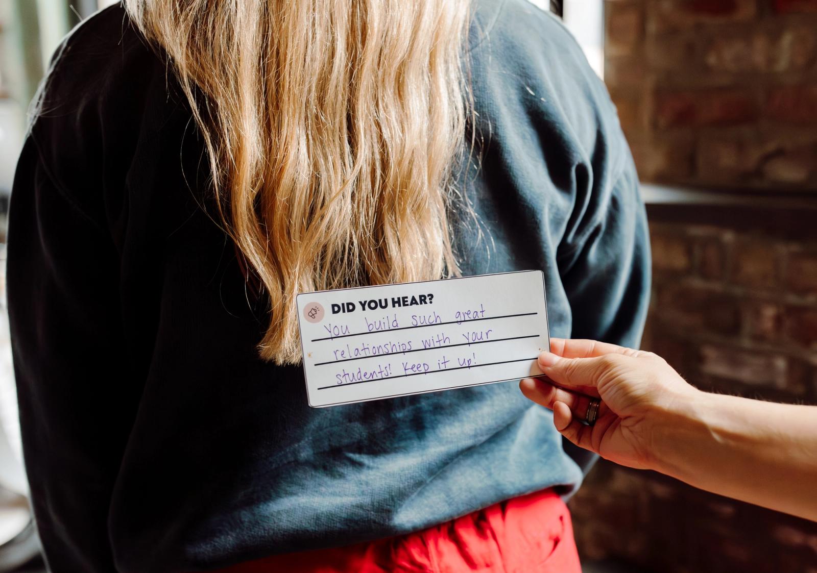 The back of a woman with long blonde hair in a black sweatshirt stands. A hand reaches in with a notecard and handwritten note on it.