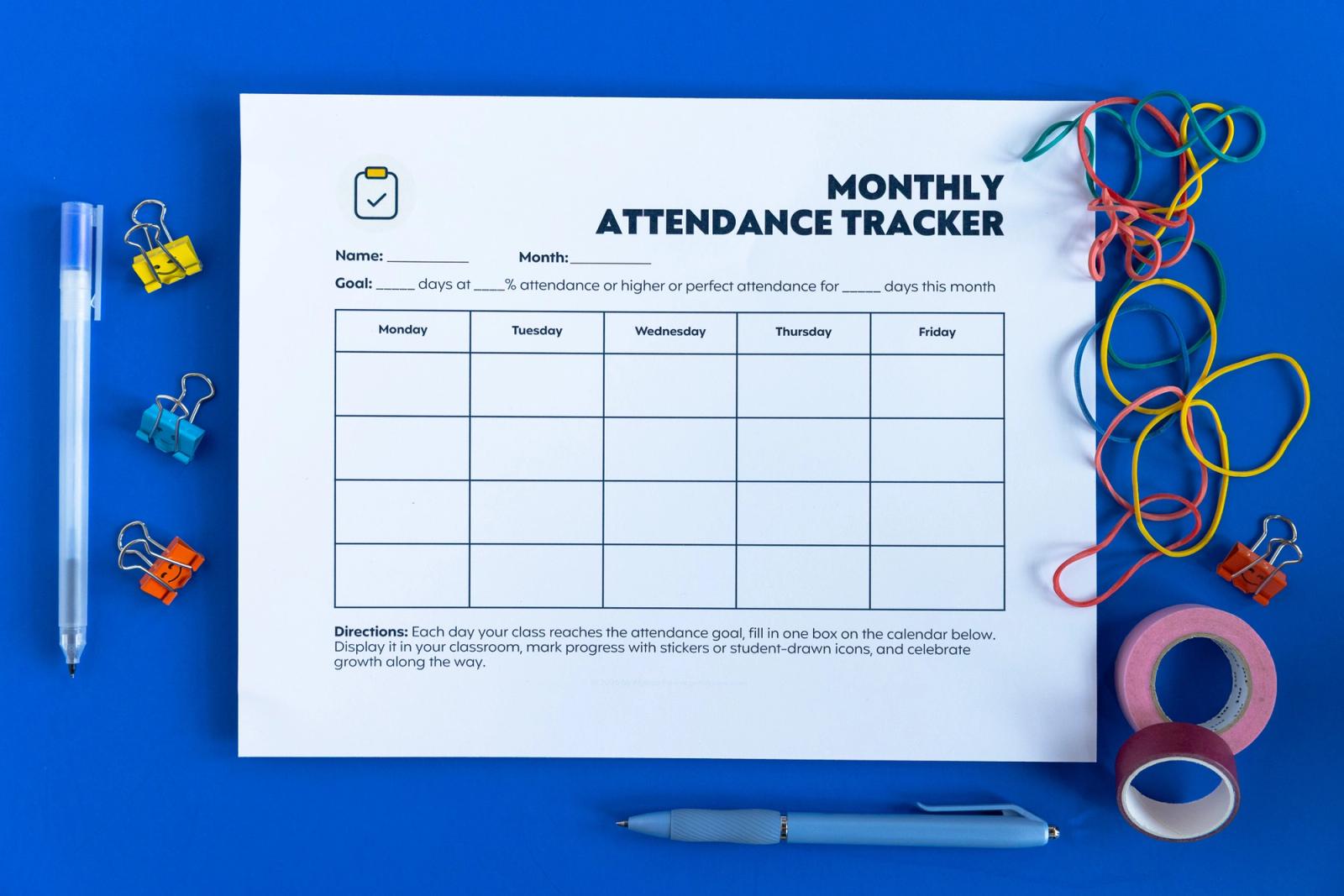 A sheet of paper that says "monthly attendance tracker" lays on a blue background. Paper clips, rubber bands, pens, and rolls of tape lay scattered around.