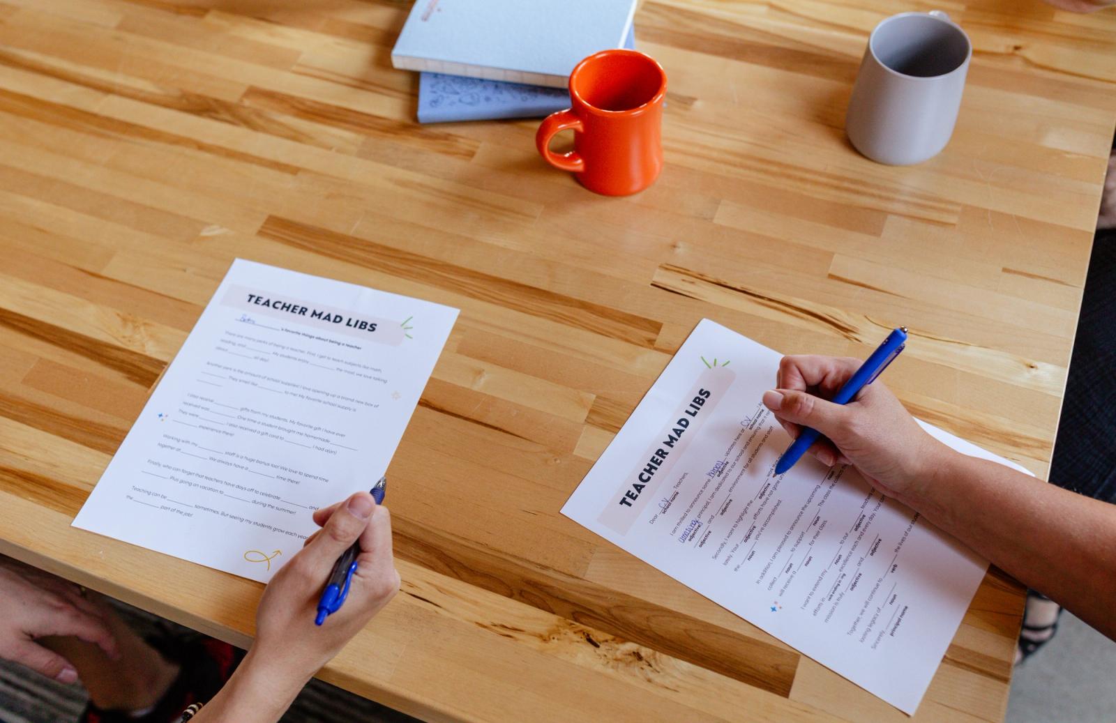 Two people sit at a wooden table writing on pieces of paper. Coffee cups and notebooks lay on the table.