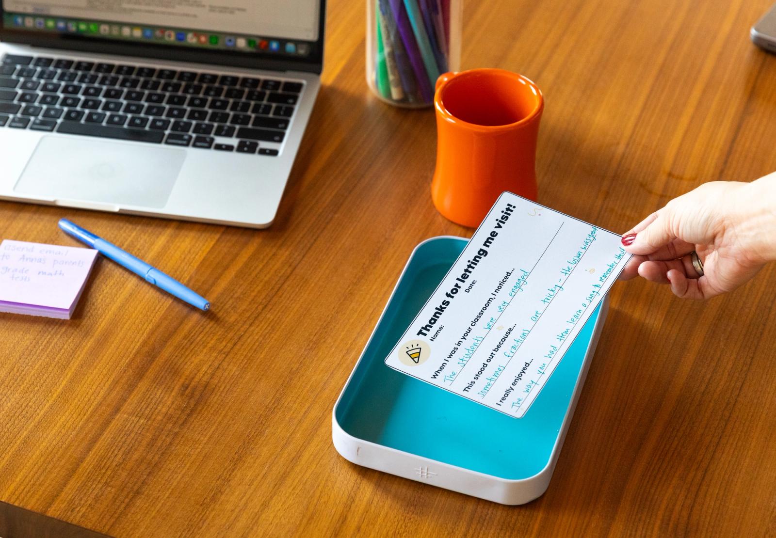 A hand drops a handwritten note in a small tray on a desk. An open laptop, coffee mug, sticky notes and pens are on the desk.