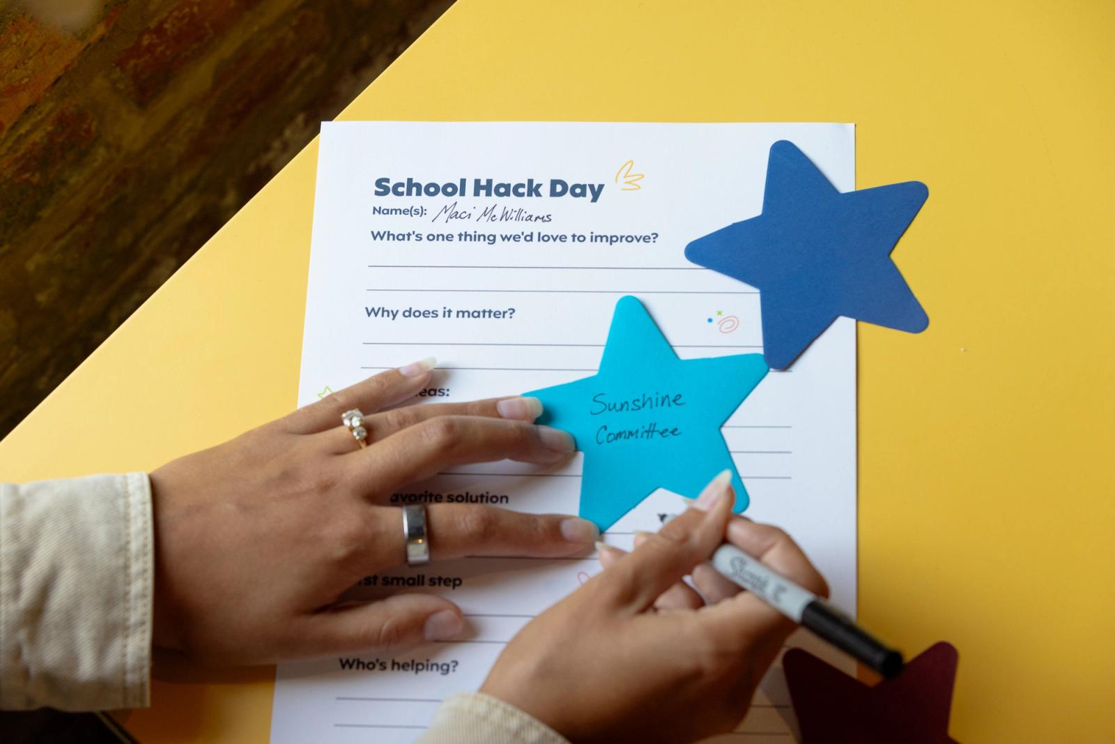 A sheet of paper that says "school hack day" lays on a yellow table. A person writes on a star next to the sheet.