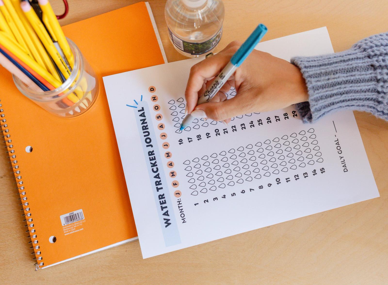 A piece of paper that says "water tracker journal" sits on a desk with an orange notebook and jar of pencils. A hand fills in the tracker.