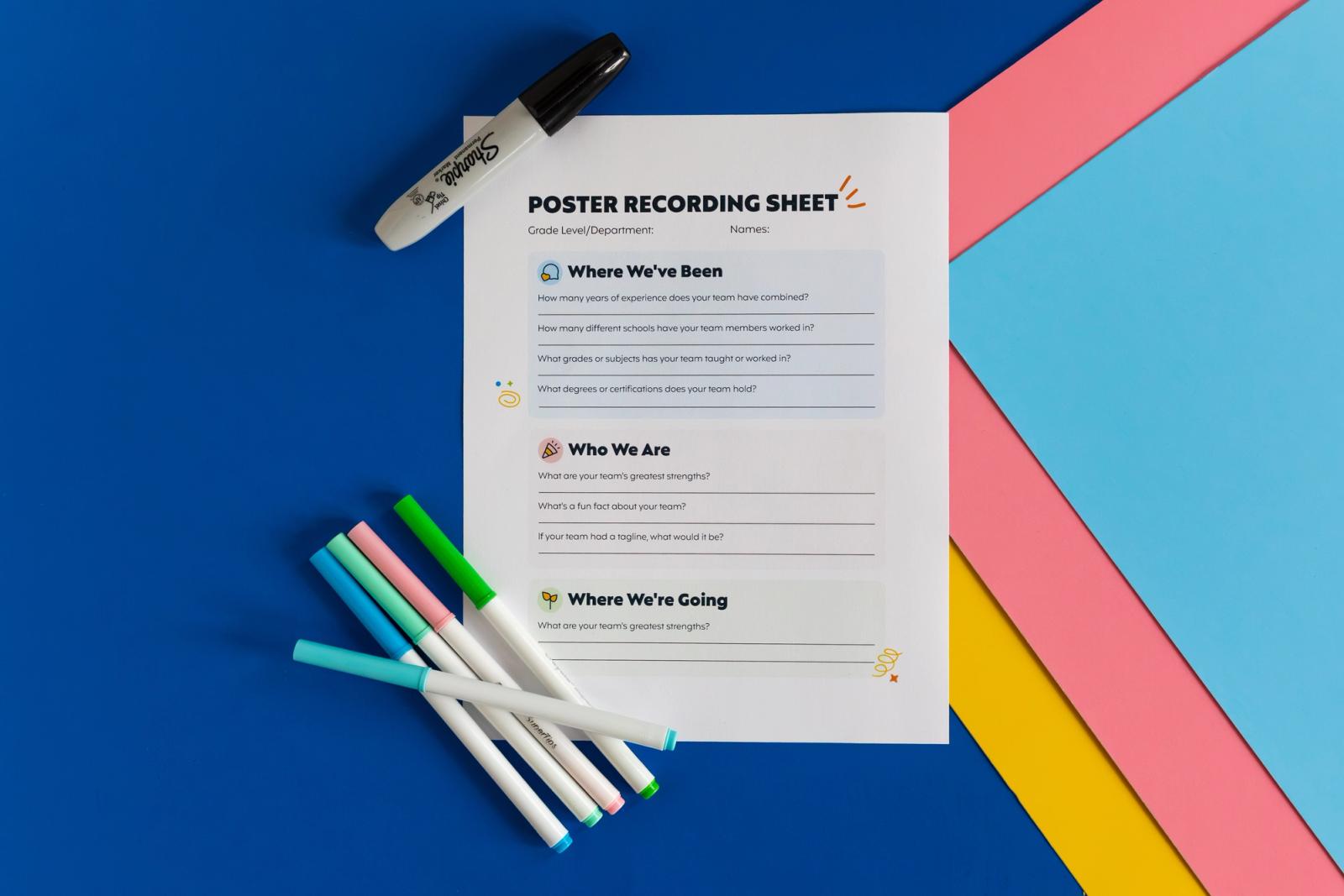 Pink, yellow, and blue poster board lay on a dark blue background. A sheet of paper and markers lay on top of them. 