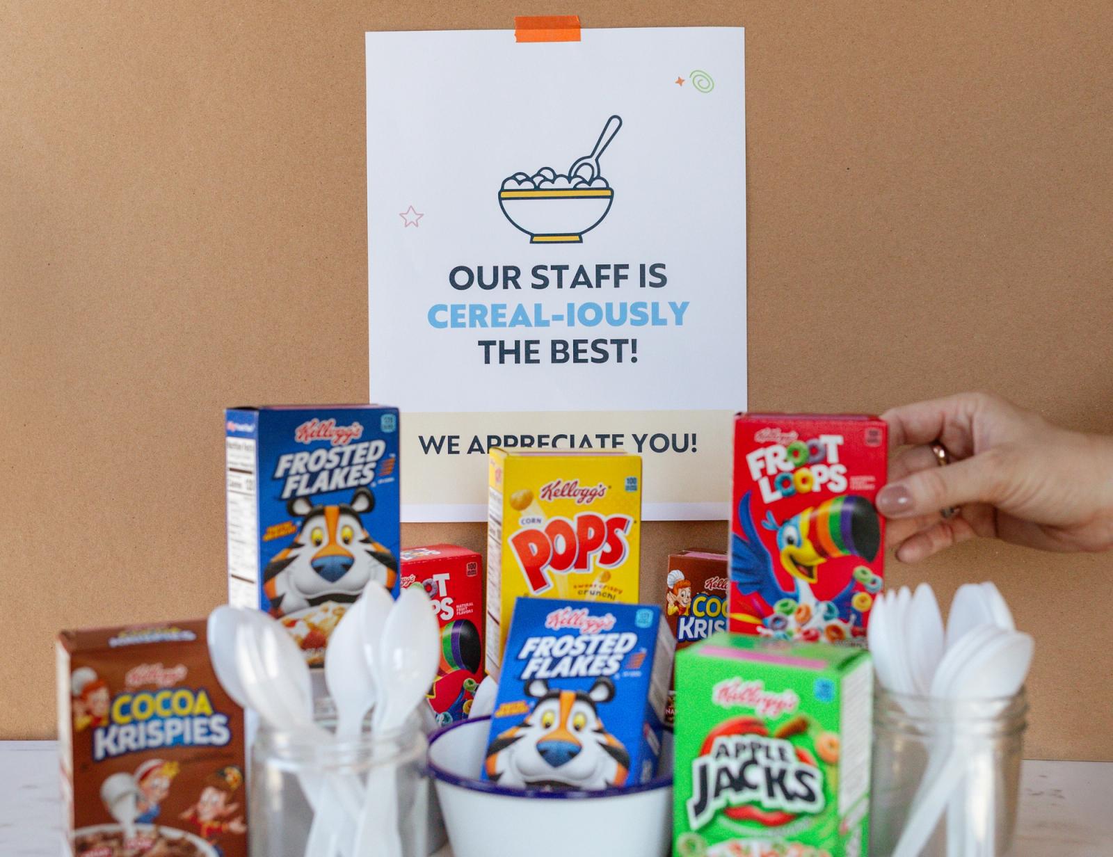 Small boxes of cereal, plastic spoons and bowls lay on a table. In the background is a brown wall with a poster that says "our staff is cereal-iously the best."