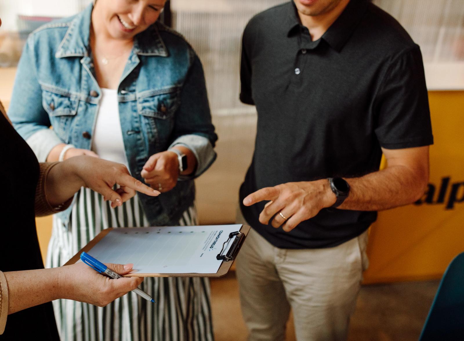 One person holds a clipboard with a piece of paper on it, and a pen in hand. Two others stand around, and all three point at the clipboard.