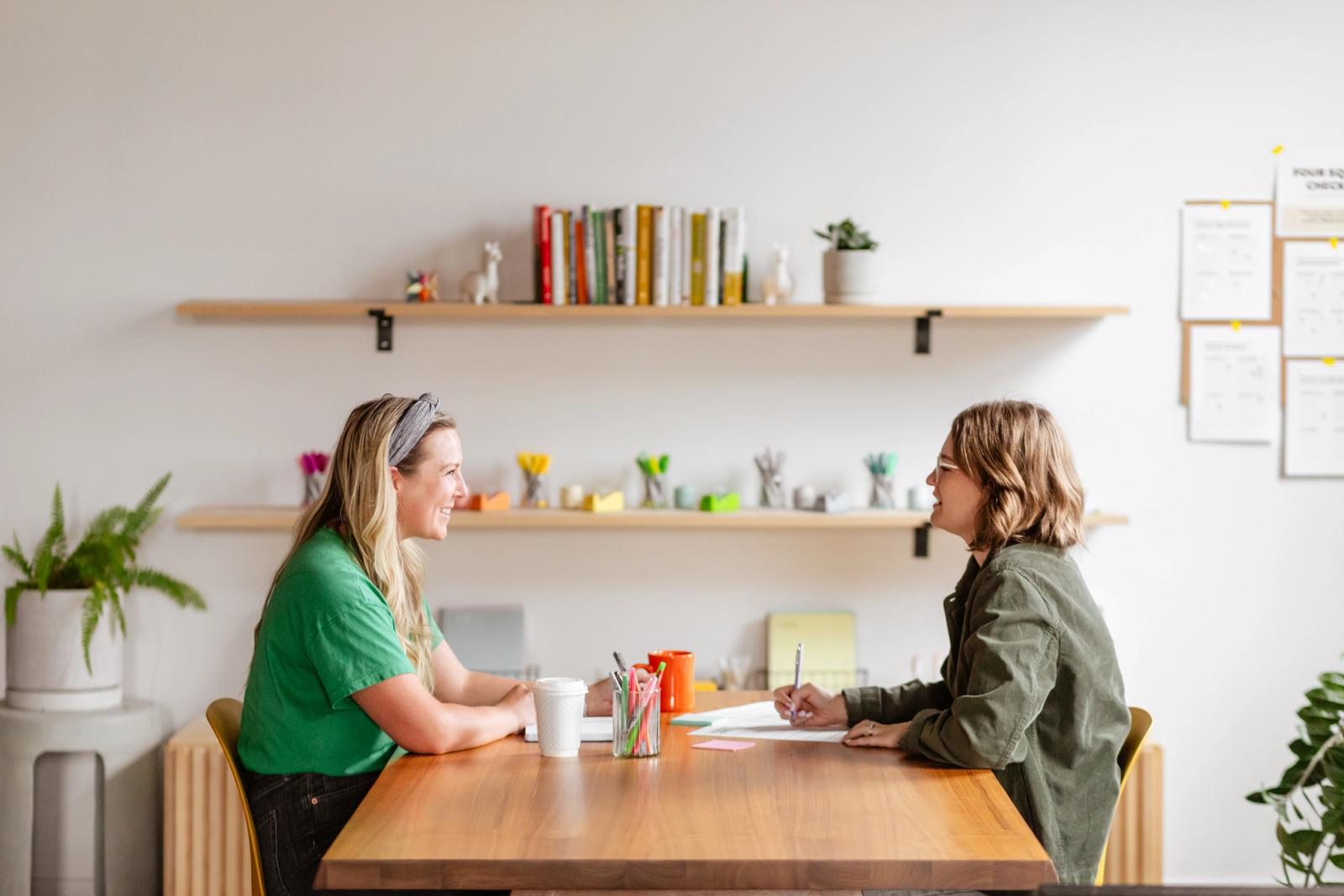 Two women sit at a table looking at each other and smiling. Pens, paper and coffee cups are on the table. There is a shelf with books and school supplies in the background.