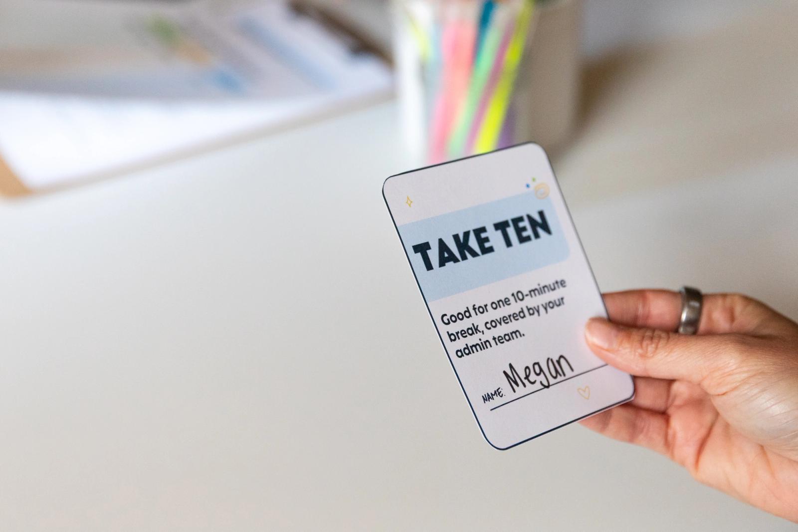 A close-up of someone holding a coupon that says "take ten" in front of a white desk with colorful pens in a jar.