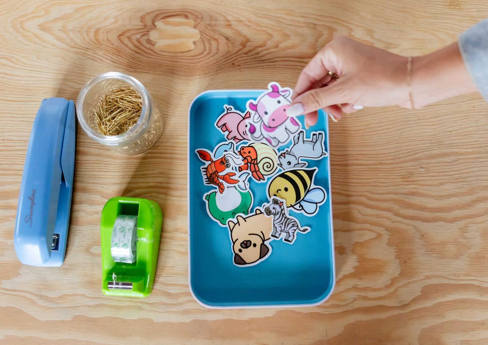 A tray with stickers in it is on a desk with school supplies around it. A hand reaches in to grab a sticker.