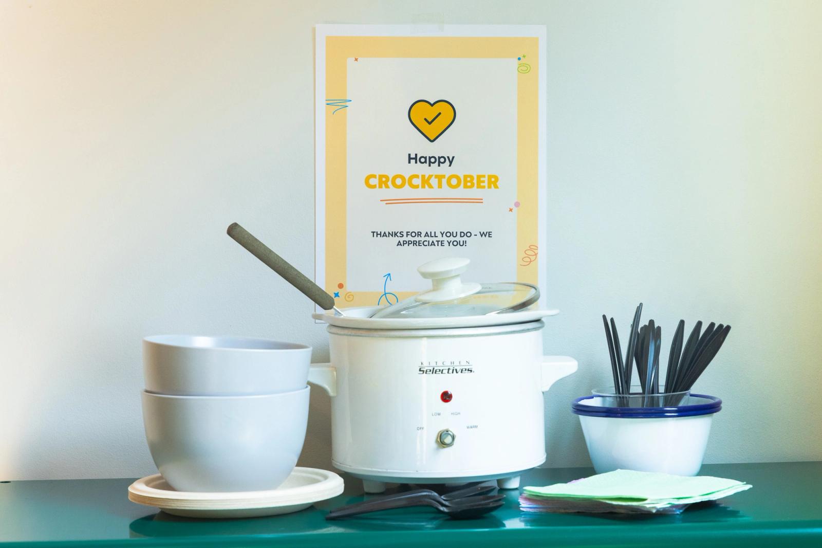 A crockpot, stack of bowls, and spoons lay on a green table. Behind it is a white wall with a poster on it.