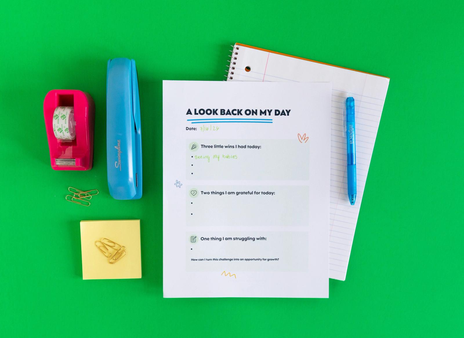 An open notebook with a piece of paper and blue pen on it lays on a green background. Next to it is a stapler, tape dispenser, sticky notes, and paper clips.