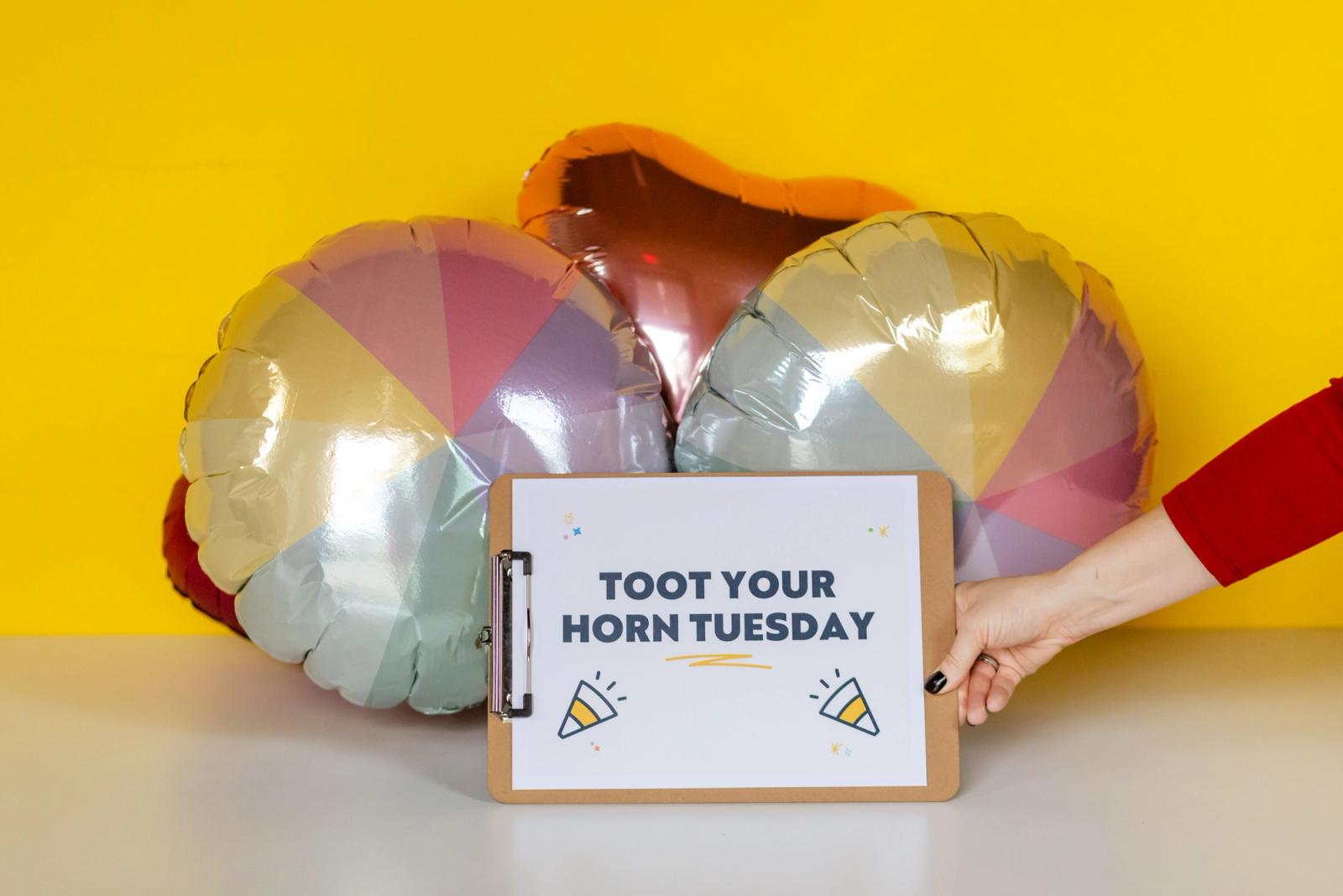 A hand holds a clipboard with a sign that says "toot your horn tuesday" on it in front of balloons.