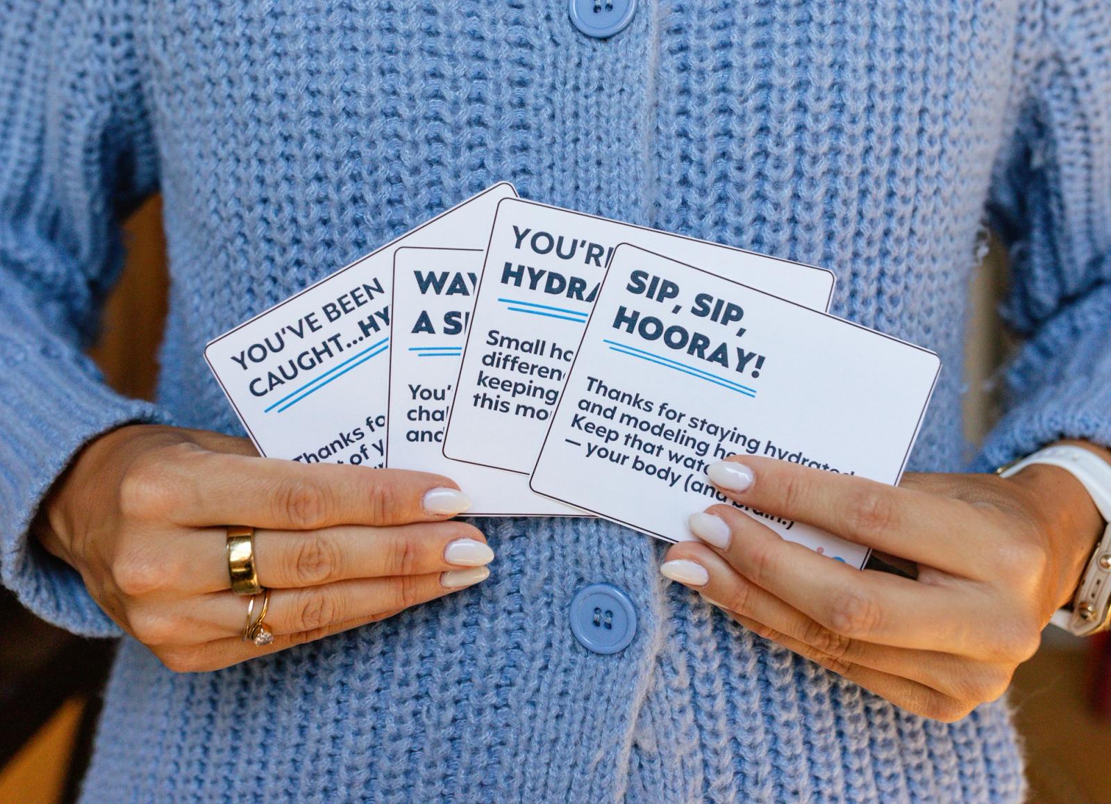Someone in a blue sweater holds small cards that say "sip, sip hooray" on them in front of them.