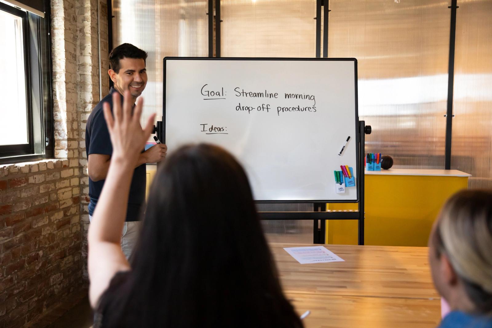 A man stands by a whiteboard with writing on it, smiling at a woman who raises her hand. 