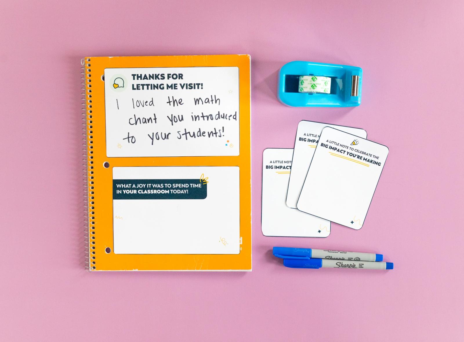 An orange notebook with two notecards on it, three small notecards, blue pens, and blue tape dispenser lay on a pink background.