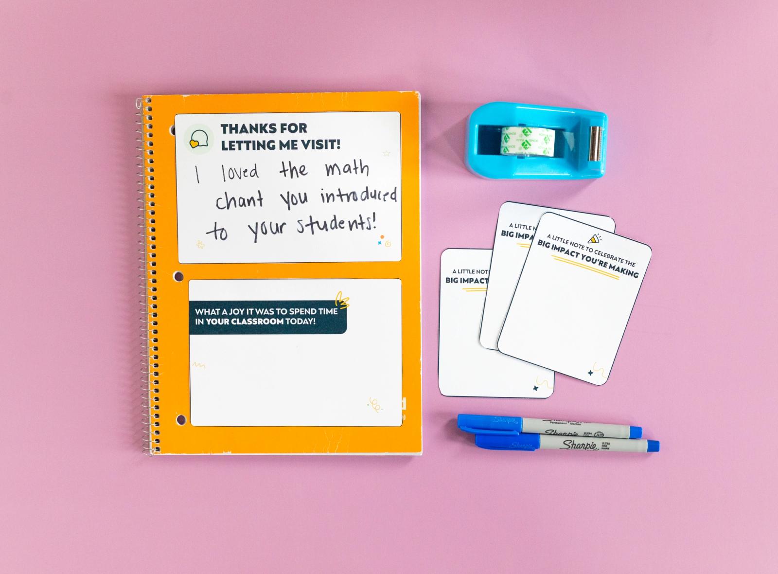 An orange notebook with two notecards on it, three small notecards, blue pens, and blue tape dispenser lay on a pink background.