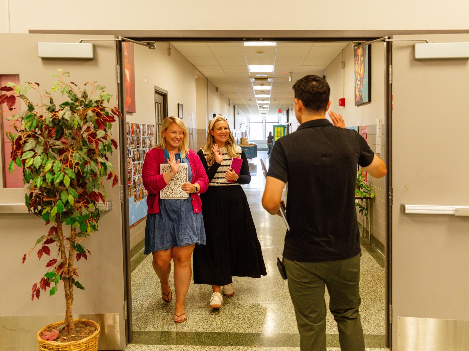 A man walks down a school hallway, and two women wave to him.