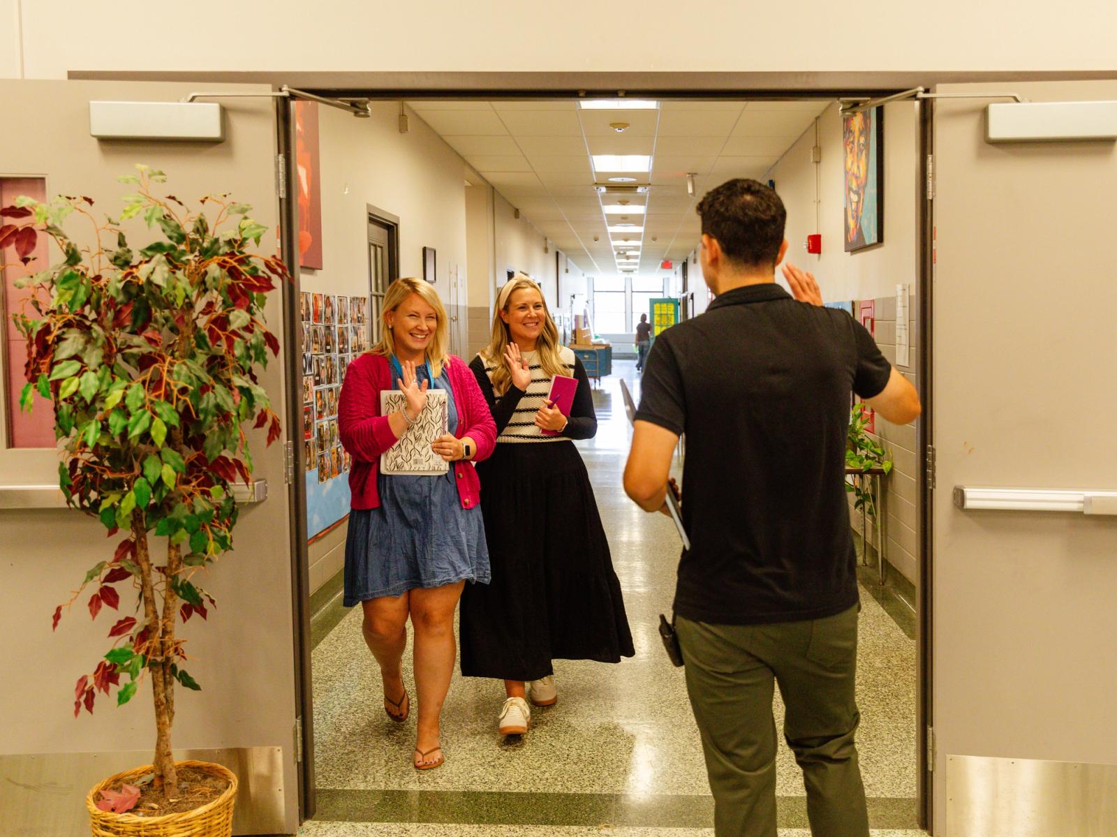 A man walks down a school hallway, and two women wave to him.