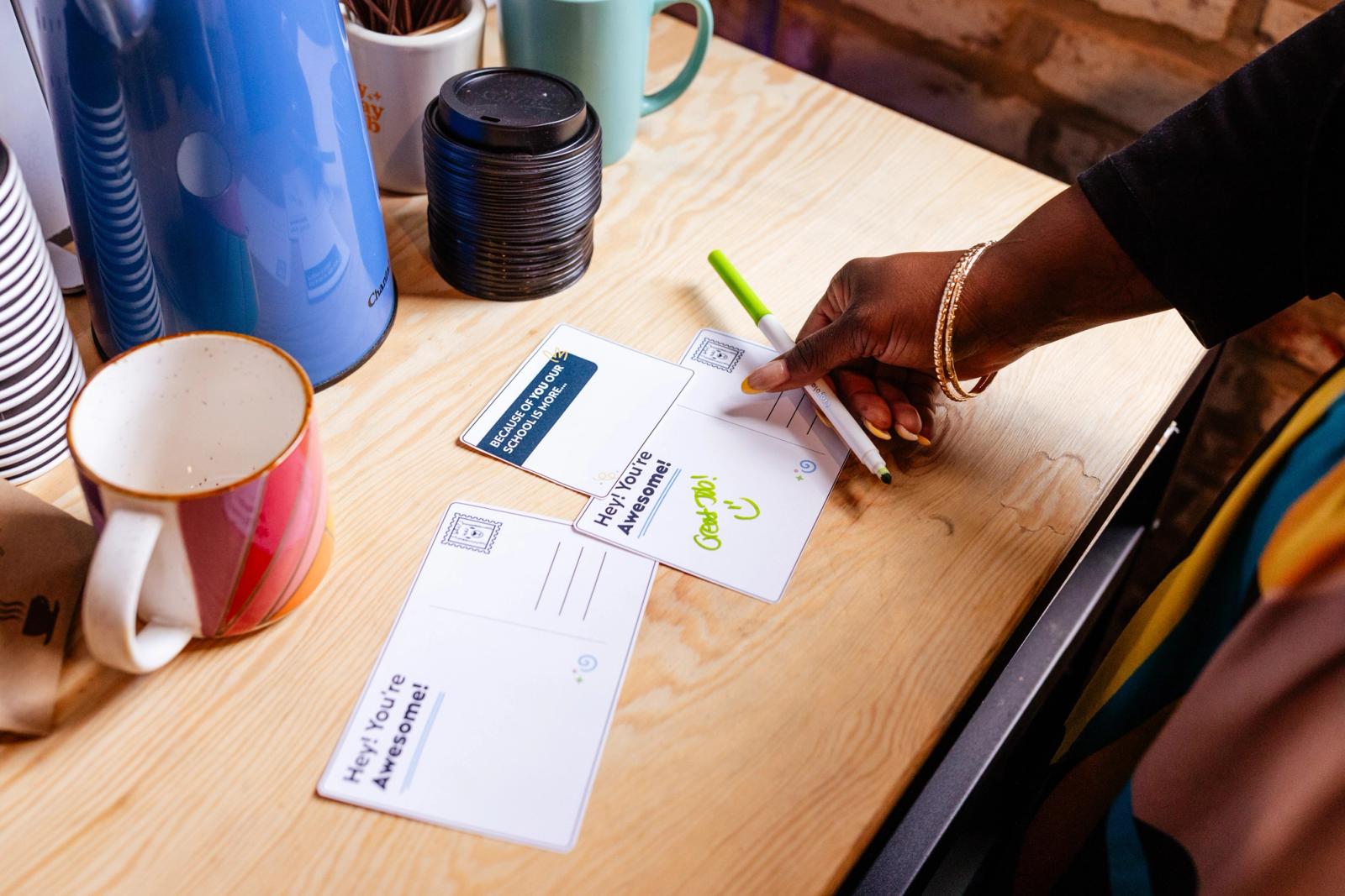 Someone writes a hand-written note on a wood table with coffee supplies on it. 