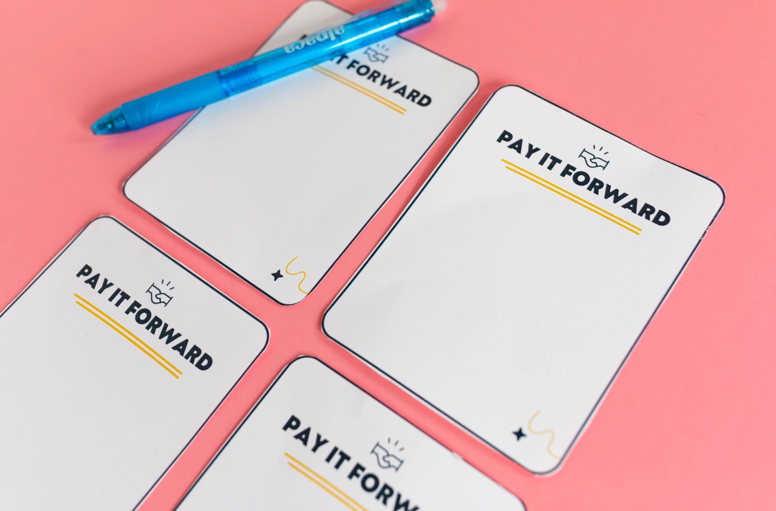 Four blank notecards that say "pay it forward" lay on a salmon background alongside a blue pen.