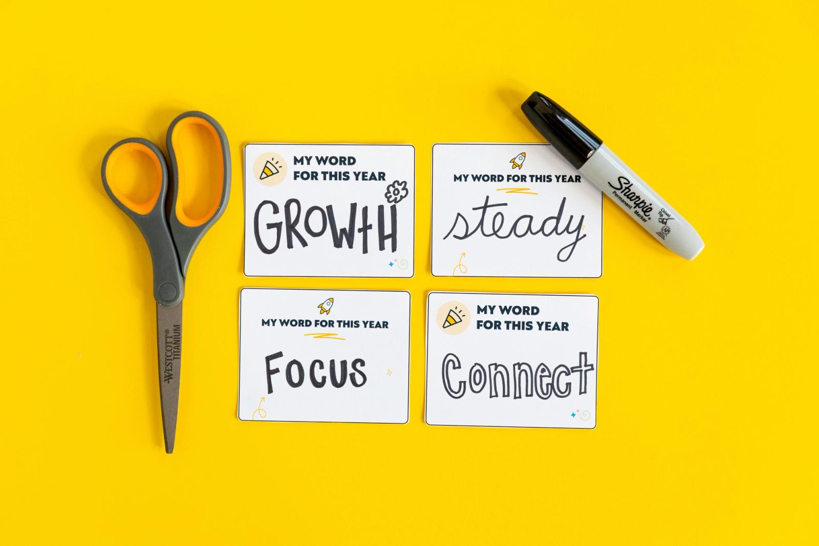 Four small notecards with one word written on each on a yellow background. A pair of scissors and a black marker lay beside the cards.