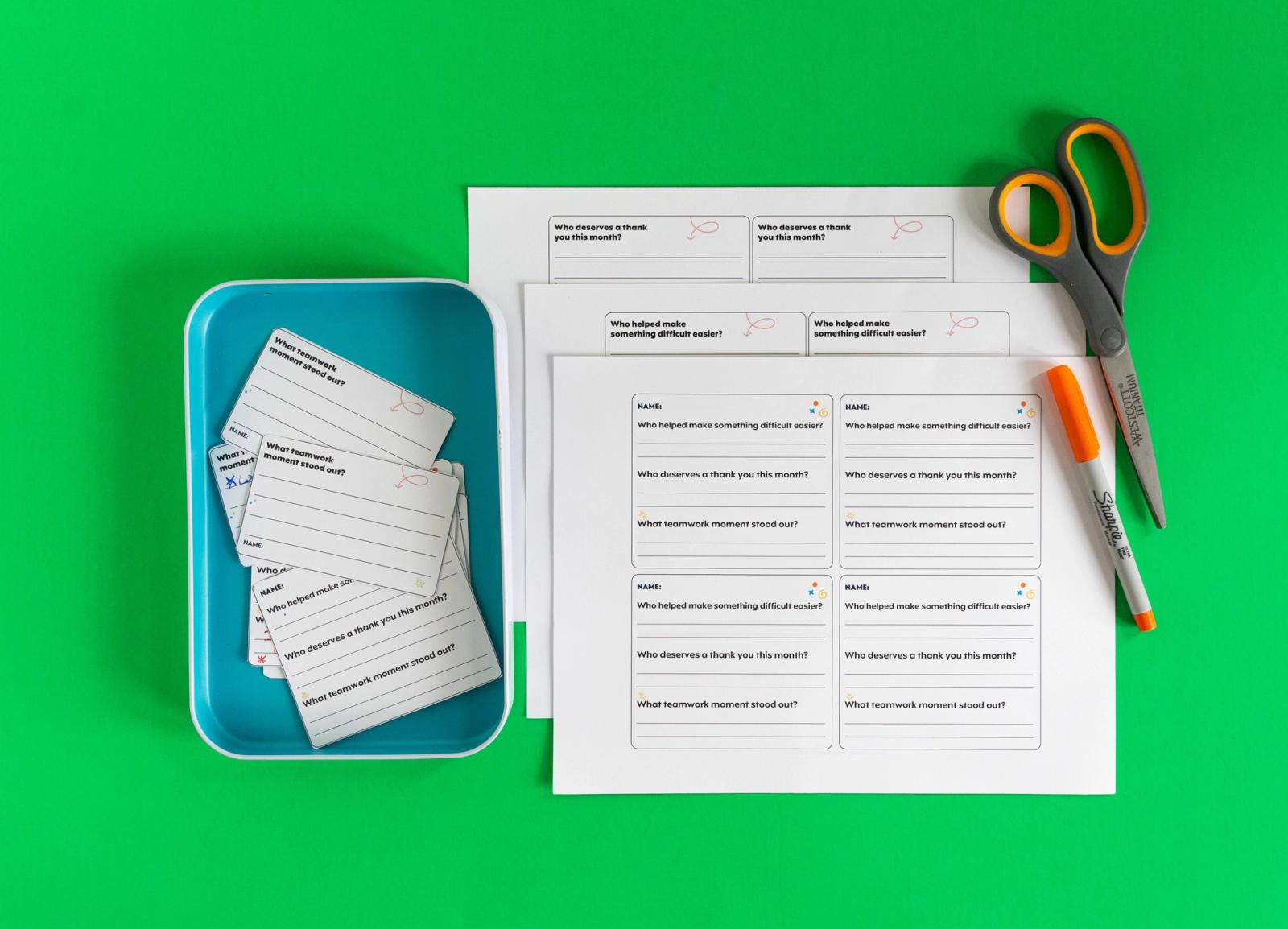 A stack of papers with notecards on them, an orange pen and pair of scissors lay on a green background. Beside them is a blue tray with more notecards in it.