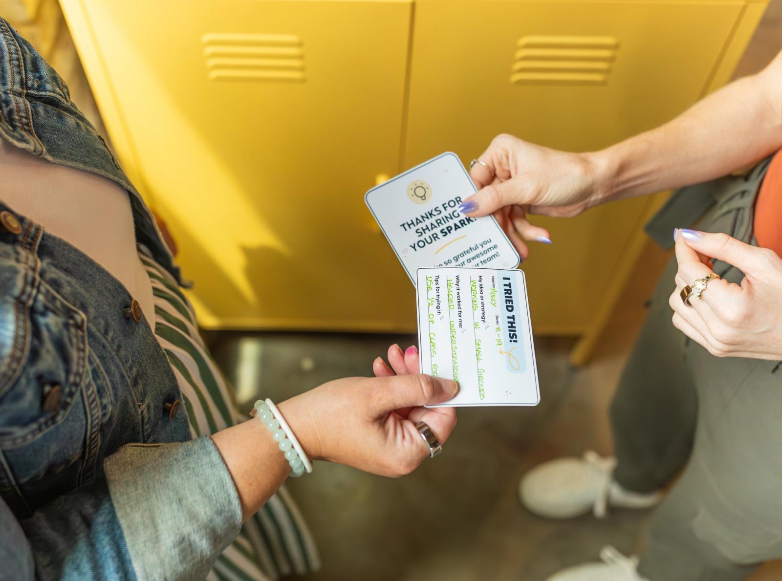 Two people stand in front of yellow lockers and their hands exchange notecards. 