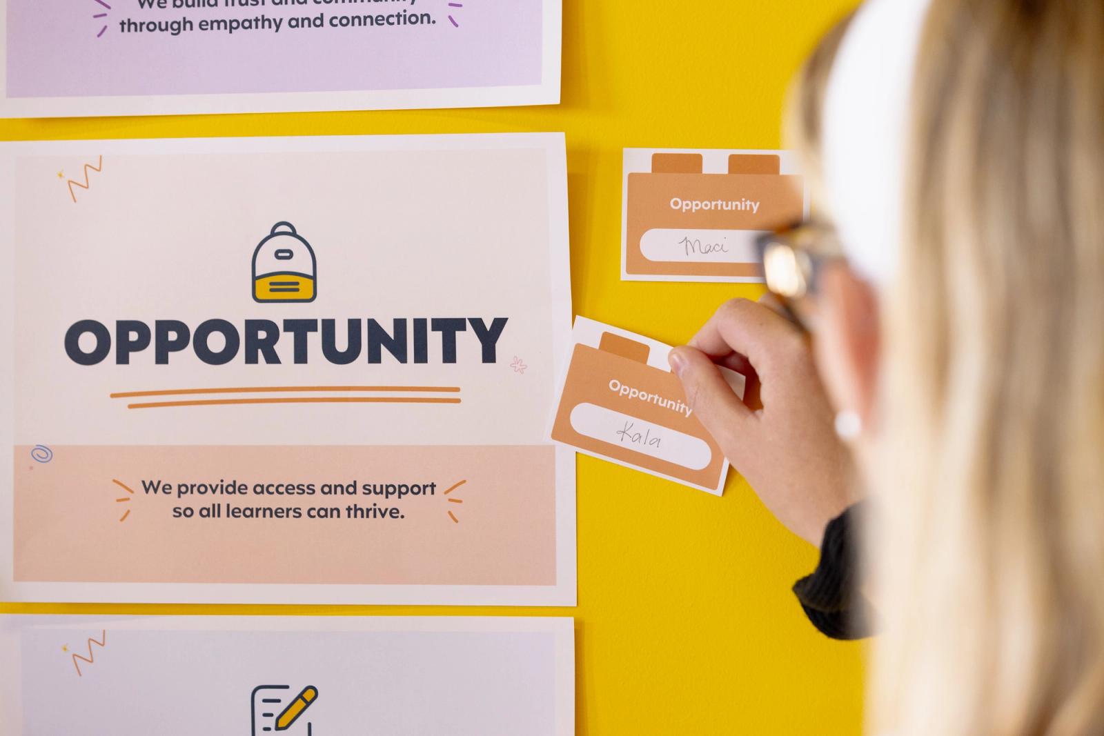 A woman places a notecard next to a poster that says "opportunity" on a yellow wall.