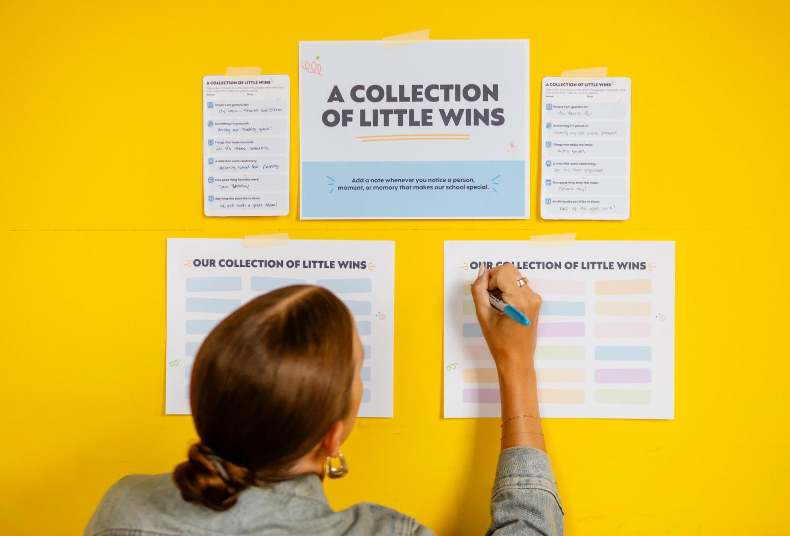 Sheets of paper are taped to a yellow wall. A woman writes on the sheet of paper. 
