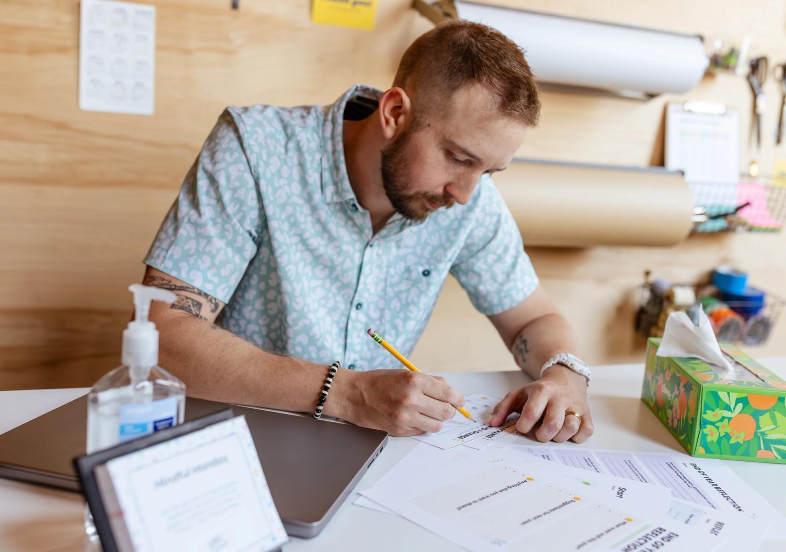 A man sits at a desk with school supplies on it. He writes with a pencil on a piece of paper. Background is a wood wall with reels of paper.
