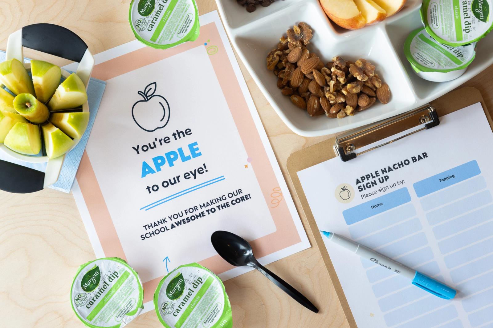 Two sheets of paper lay on a wood table. Around them are apple slices, jars of caramel sauce, and a tray of nuts.