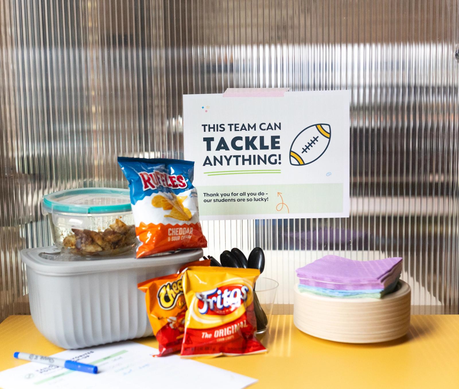 A football themed poster hangs on a glass wall in front of a yellow table with snacks, paper plates, and napkins.