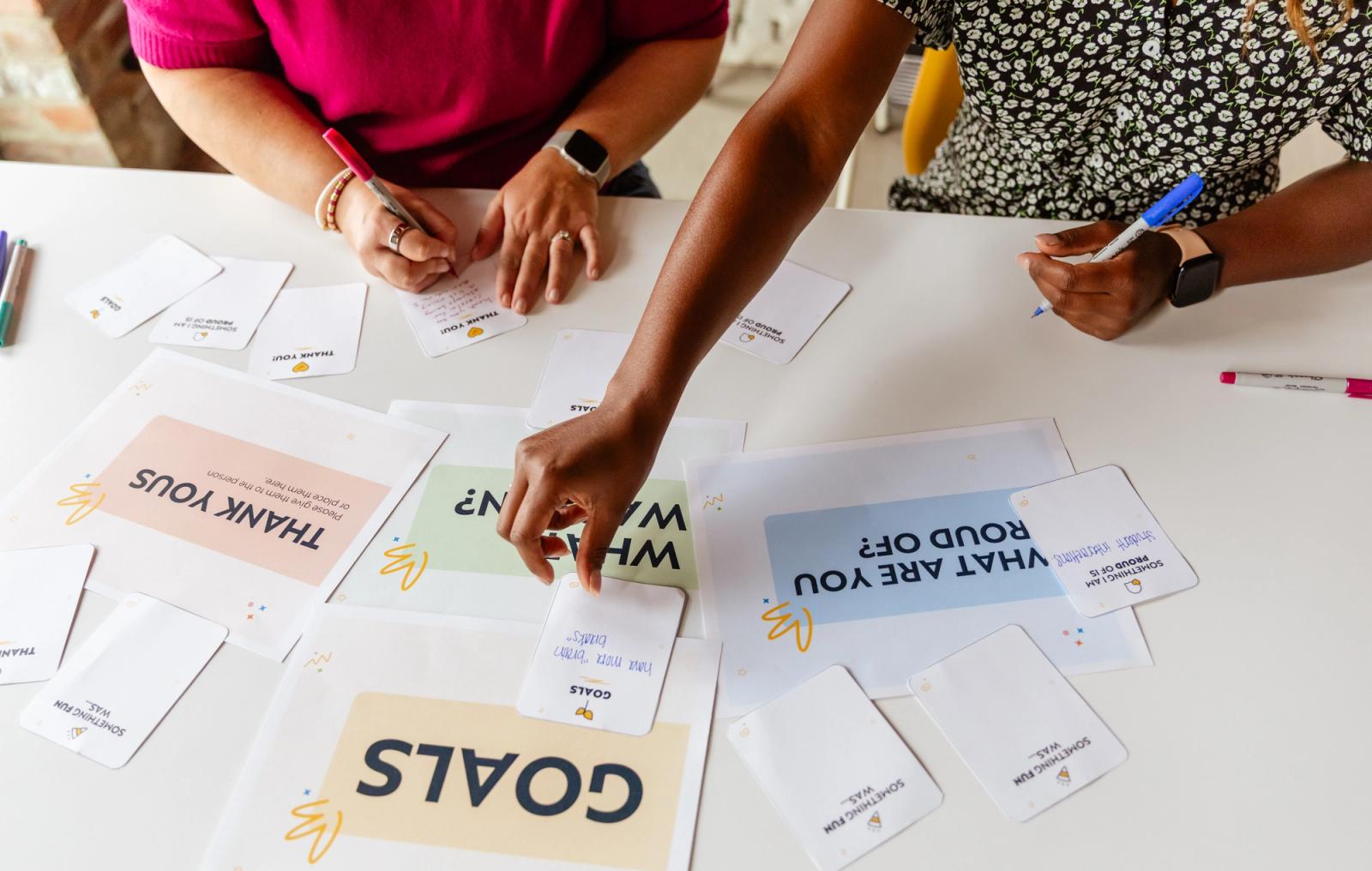 Two women sit at a table with pens in hands. they write on notecards.