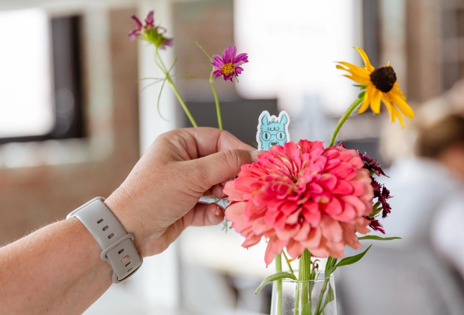 A hand places a small alpaca sticker in a vase of flowers.