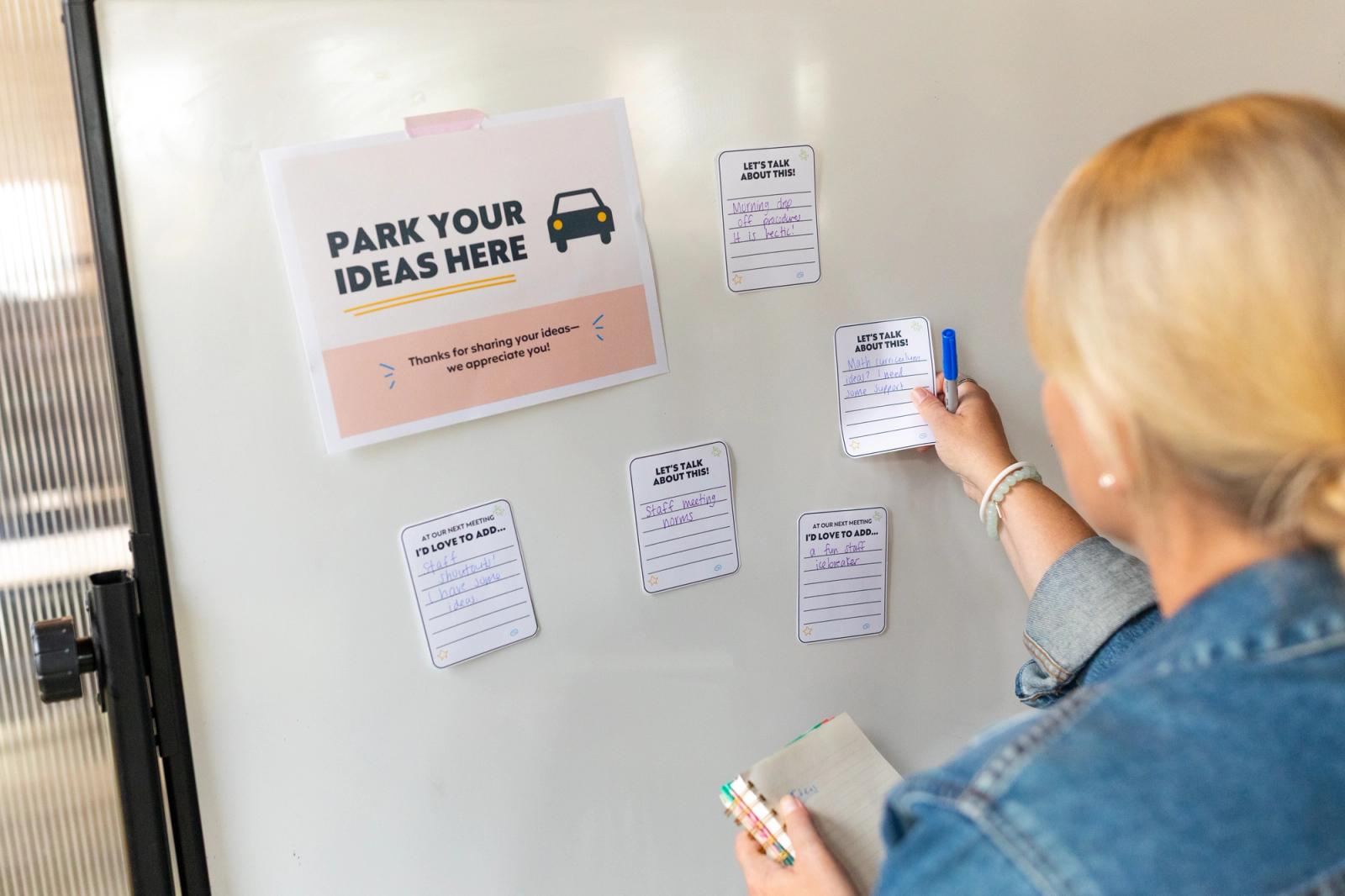 A woman stands in front of a whiteboard that has a poster on it. She tapes a notecard near the poster.