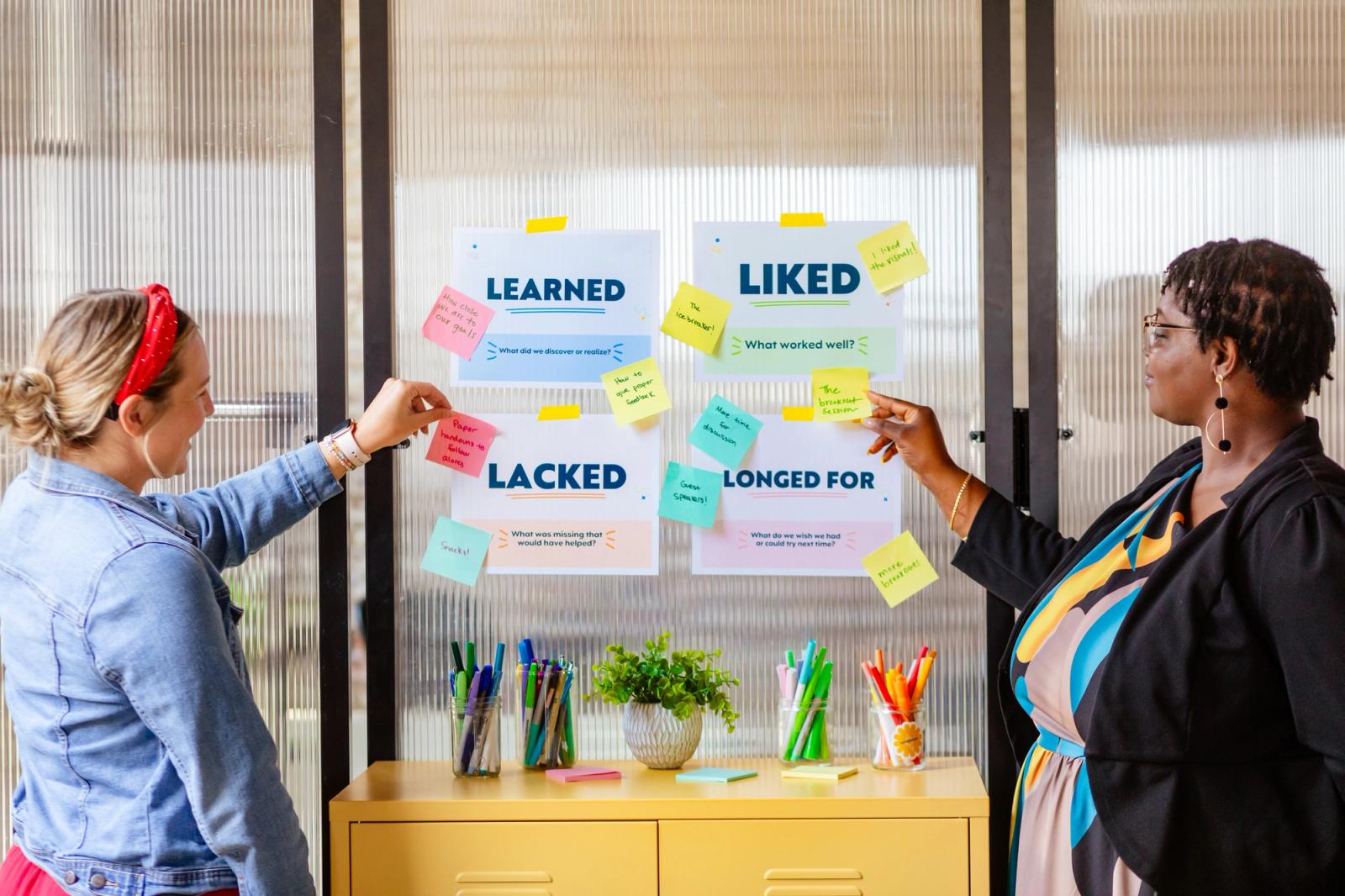 Two women place sticky-notes under four posters with "learned, liked, lacked, and longed for" printed on them on a frosted glass wall. A yellow table with pens and a plant is between them.