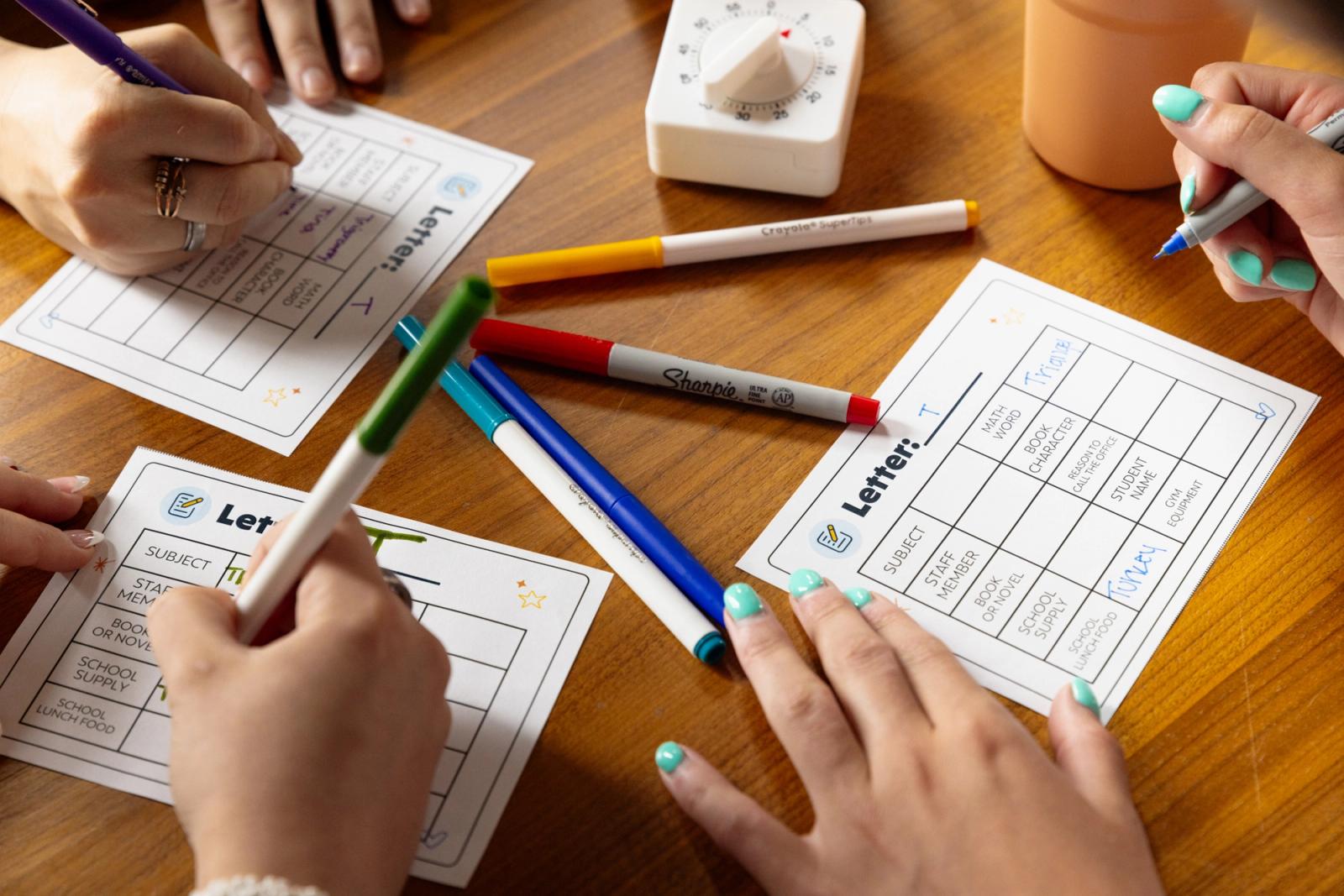 Hands write with markers on pieces of paper laying on a wood table. A timer sits in the background.