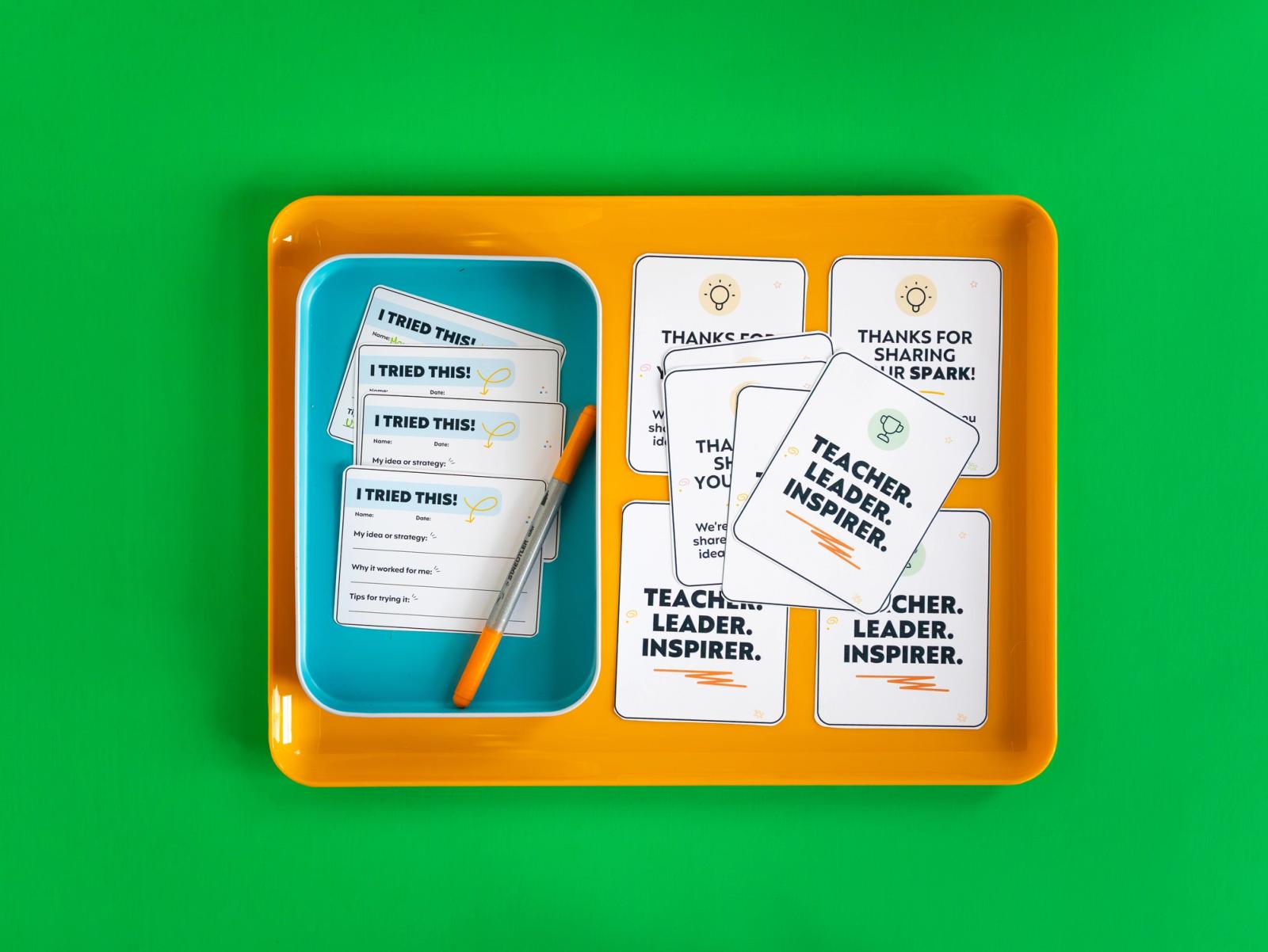 An orange lunch tray sits on a green background. In the lunch tray is a small blue container with notecards and an orange pen in it. Next to the tray are more notecards.
