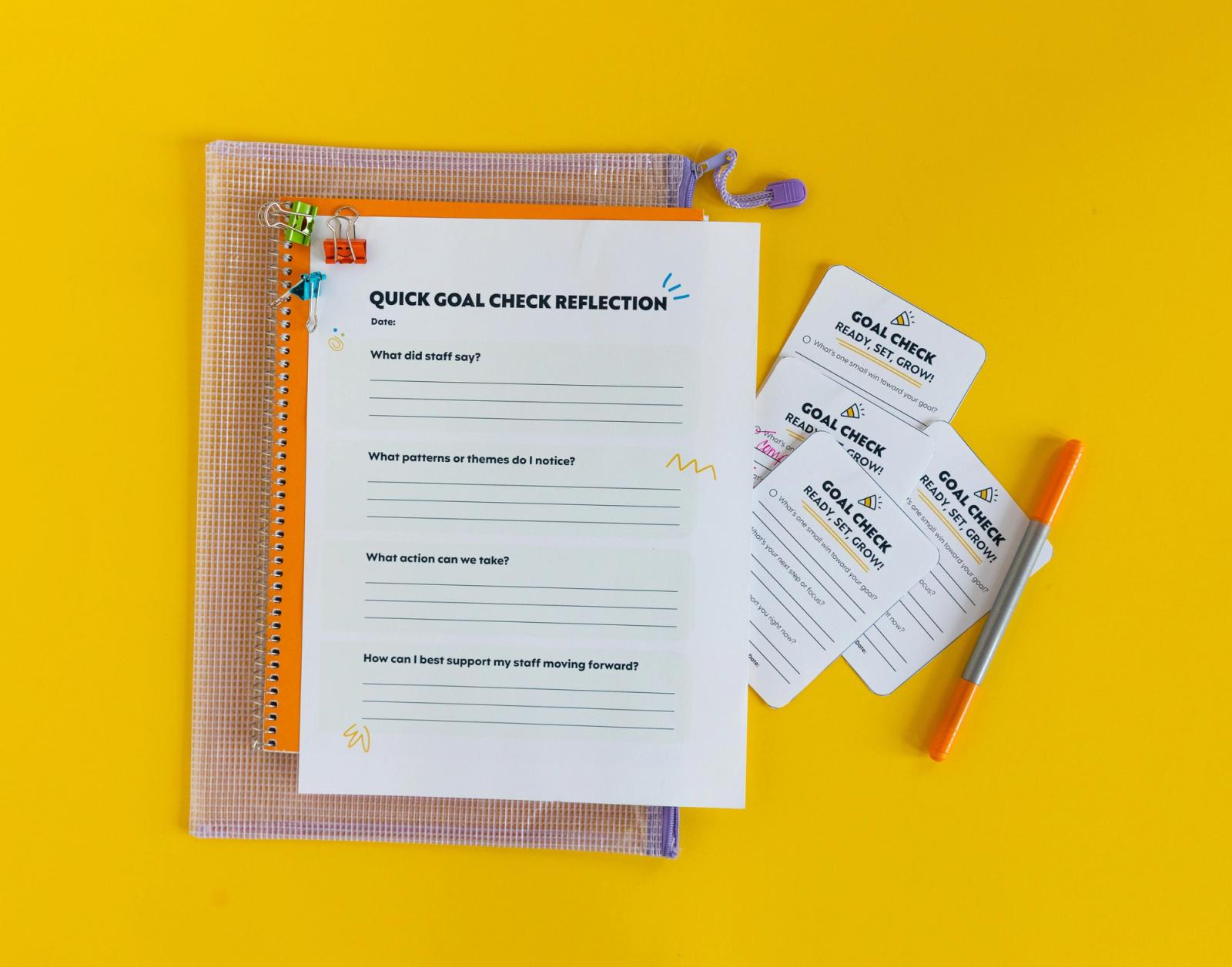 A plastic bag, orange notebook, and paper that says "quick goal check reflection" lay on a yellow background. Small notecards and an orange pen lay next to them.