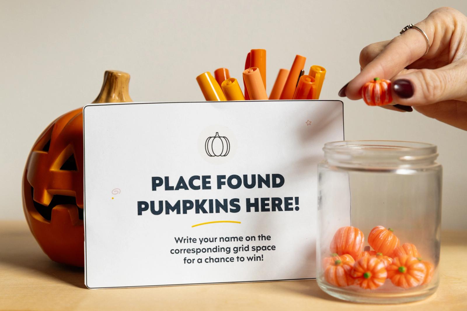 A paper that says "place found pumpkins here" sits on a table with a pumpkin next to it. A hand places small pumpkins in a jar.