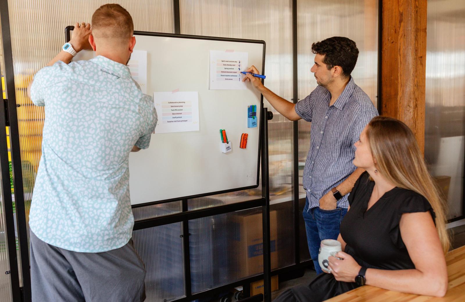 Two men stand in front of a whiteboard and write on pieces of paper taped to the board. A woman sits at the table with a cup of coffee watching them.