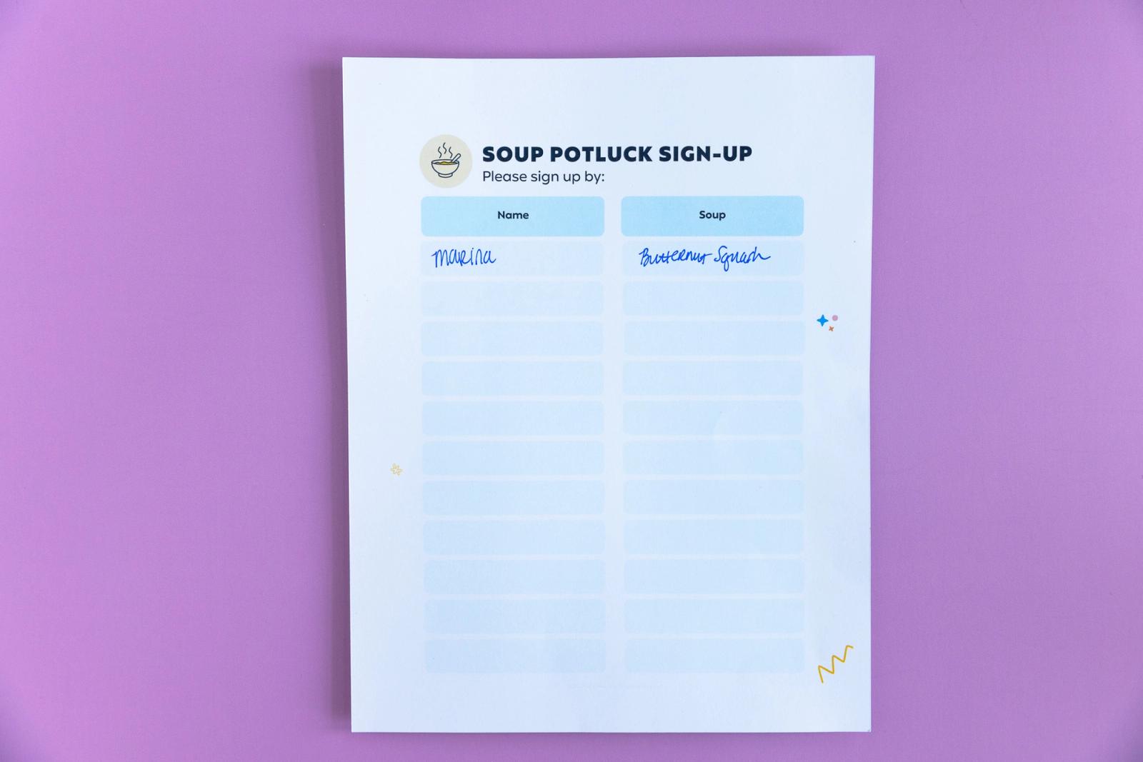 A sheet of paper that says "soup potluck sign-up" lays on a light purple background.