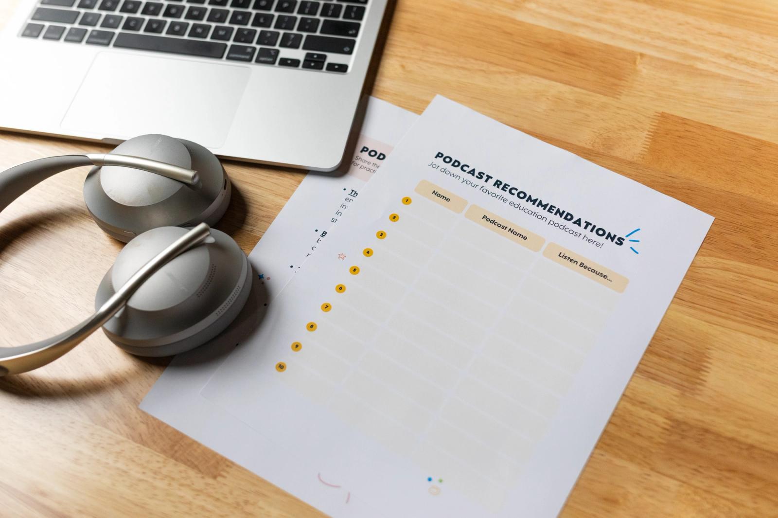 A laptop, set of gray over-the-ear headphones and two sheets of paper lay on a wood table.
