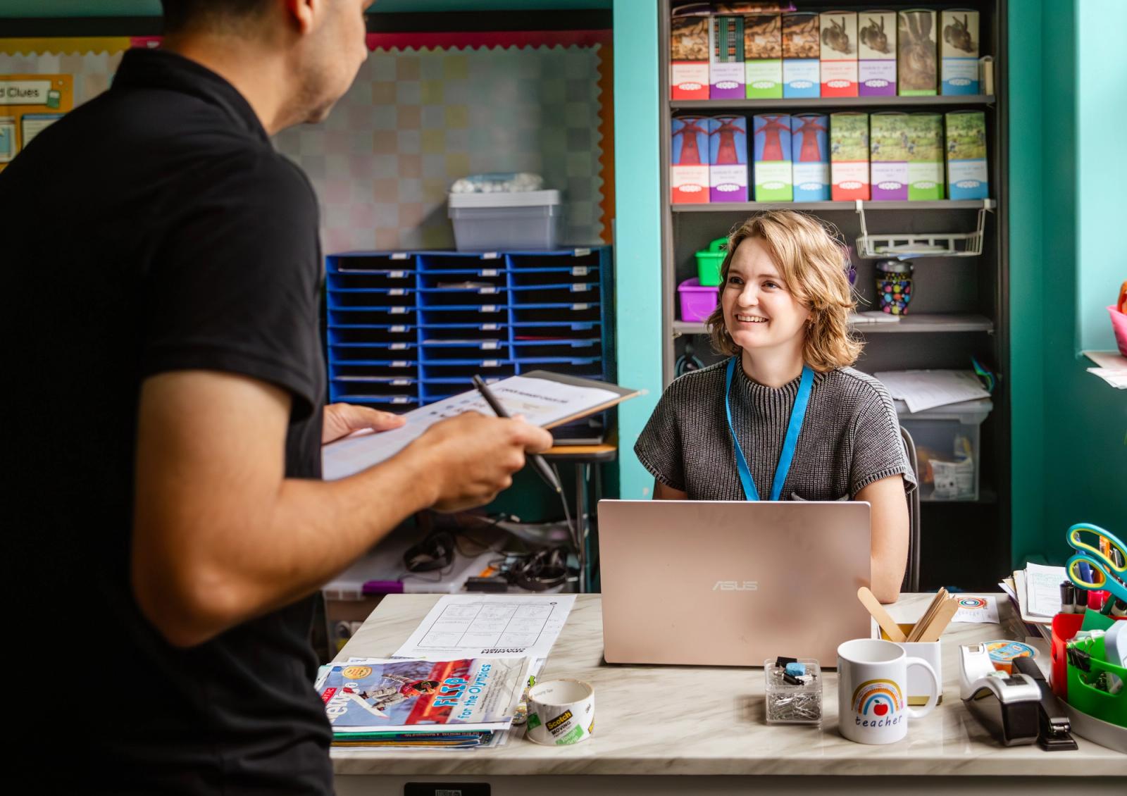 A man holding a clipboard stands in front of a woman seated at a desk in a classroom.