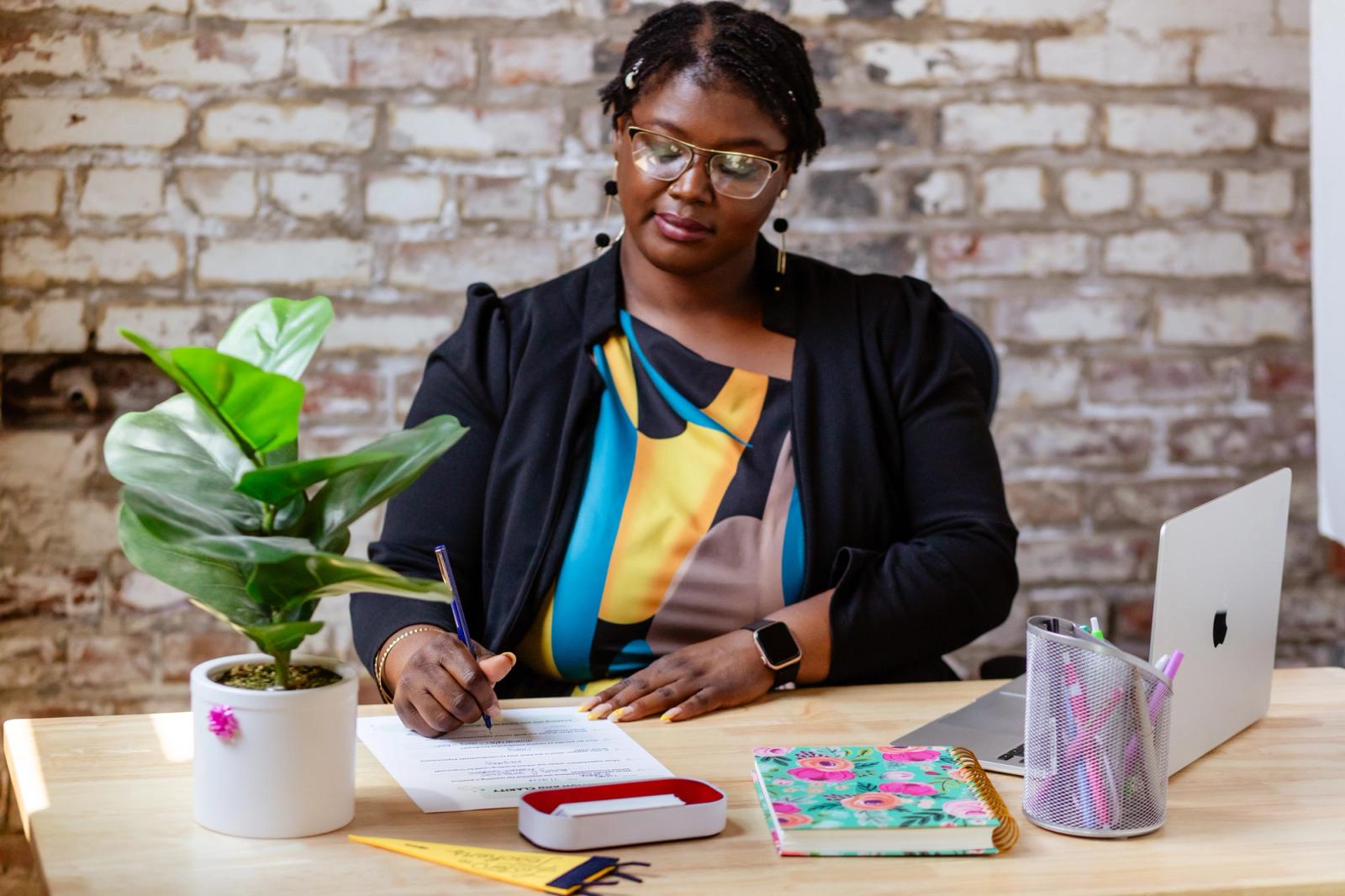A woman sits at a wooden desk with a laptop, plant, and writing supplies on it. She writes on a piece of paper in front of a brick background.